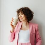 Confident woman in pink suit smiling with coffee cup indoors against white wall.