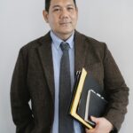Positive Asian male office worker in formal suit with paper folders in hand standing with hand in pocket against white wall